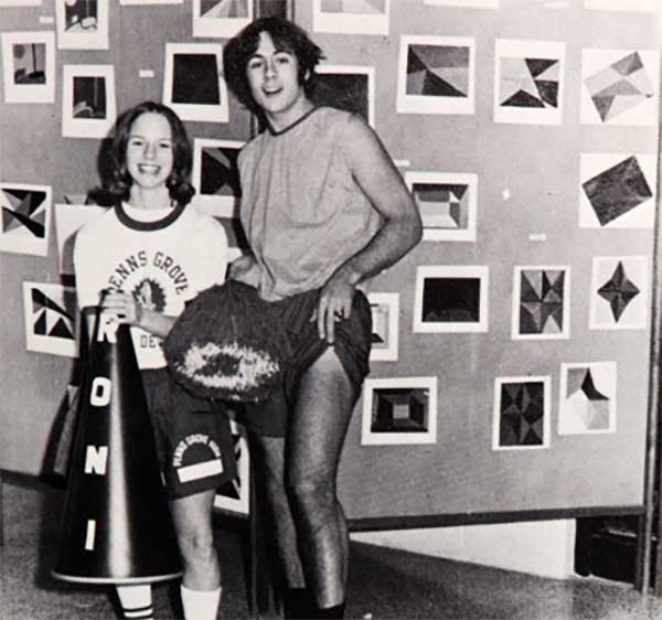 A young woman in a cheerleader outfit holding a megaphone stands next to a young man in athletic clothes in front of a wall covered with geometric artwork. Both are smiling at the camera.