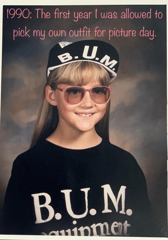 Young child with long blond hair and pink sunglasses, wearing a B.U.M. Equipment shirt and matching cap, smiles for a school photo. Text above reads: “1990: The first year I was allowed to pick my own outfit for picture day.”