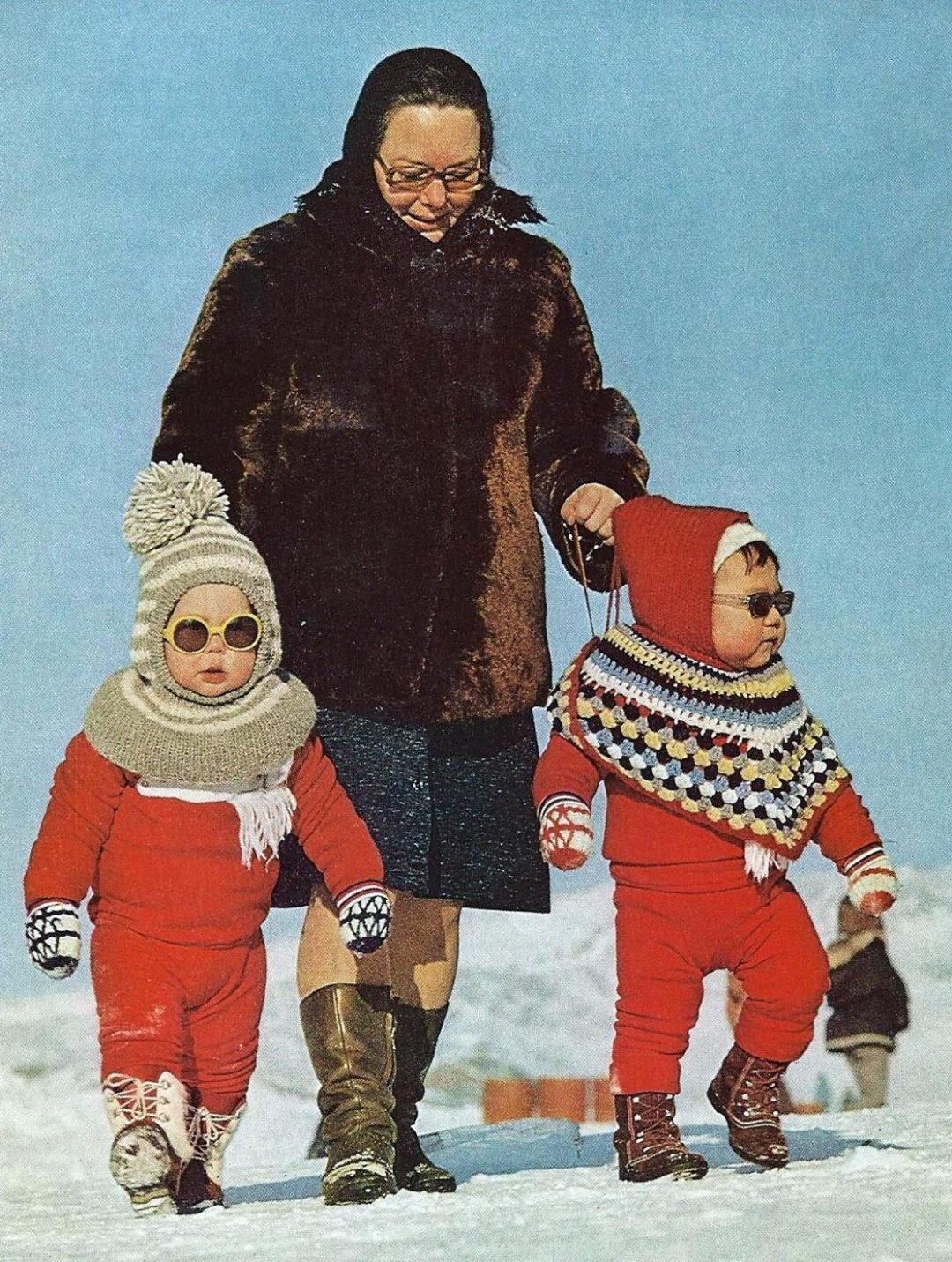A woman in a dark coat and boots walks with two young children in red winter outfits and sunglasses across a snowy landscape. The children wear matching hats and scarves, and one has a colorful poncho.
