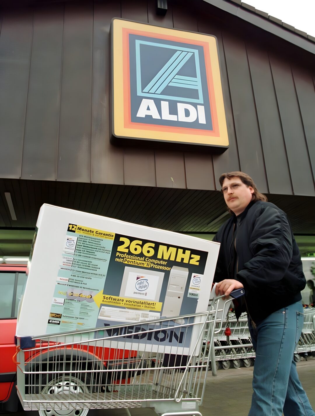 A person stands outside an ALDI store, pushing a shopping cart with a boxed 266 MHz computer. The large ALDI sign is visible above the entrance in the background.