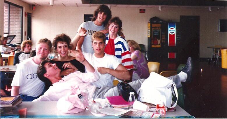 A group of six young people pose playfully around a cafeteria table, smiling and laughing. Magazines and bags are on the table, and vending machines are seen in the background.