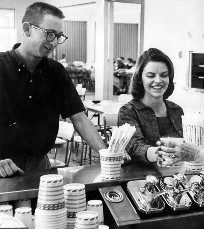 A young man and woman stand at an ice cream or soda counter. The woman smiles while receiving a drink, and the man stands beside her. Paper cups and drink dispensers are visible on the counter.