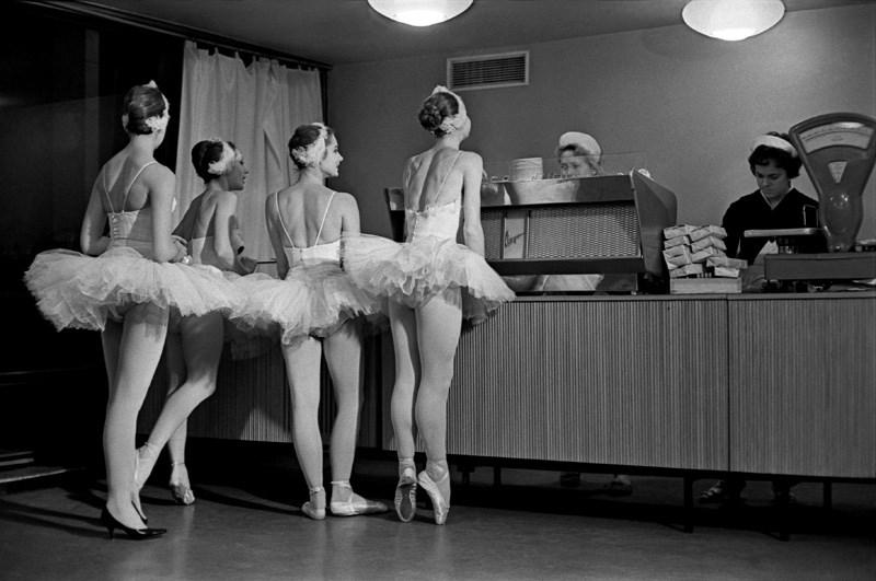 Four ballerinas in tutus and pointe shoes stand at a counter, being served by two women in uniforms, in what appears to be a cafeteria or canteen.