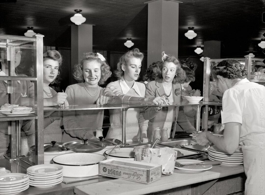 Four young women stand in line at a cafeteria counter, waiting to be served by a worker in a white uniform, with plates and food trays visible in the foreground.