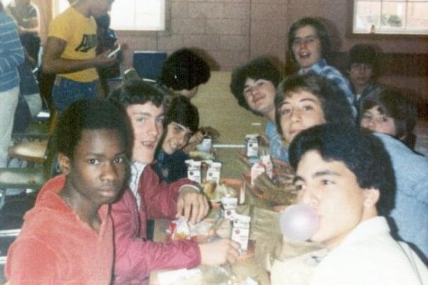 A group of teenagers sit around a cafeteria table eating lunch. Several milk cartons and food wrappers are on the table. One person blows a bubble with gum while others smile at the camera. The room appears casual and lively.