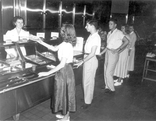 A black-and-white photo shows young people in 1950s-style clothing standing in line at a cafeteria counter, being served food by a woman behind the counter.