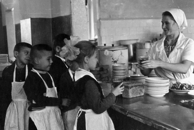 Four children wearing aprons stand in line at a cafeteria counter, receiving food from a woman in a white uniform and headscarf who is handing a jar to the girl at the front of the line.