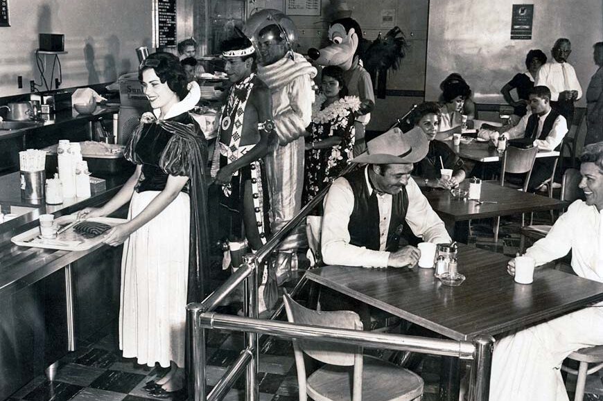 A vintage photo of people in a cafeteria; some are dressed as classic Disney characters such as Snow White and Goofy, while others wear casual or cowboy attire, sitting at tables and waiting in line for food.