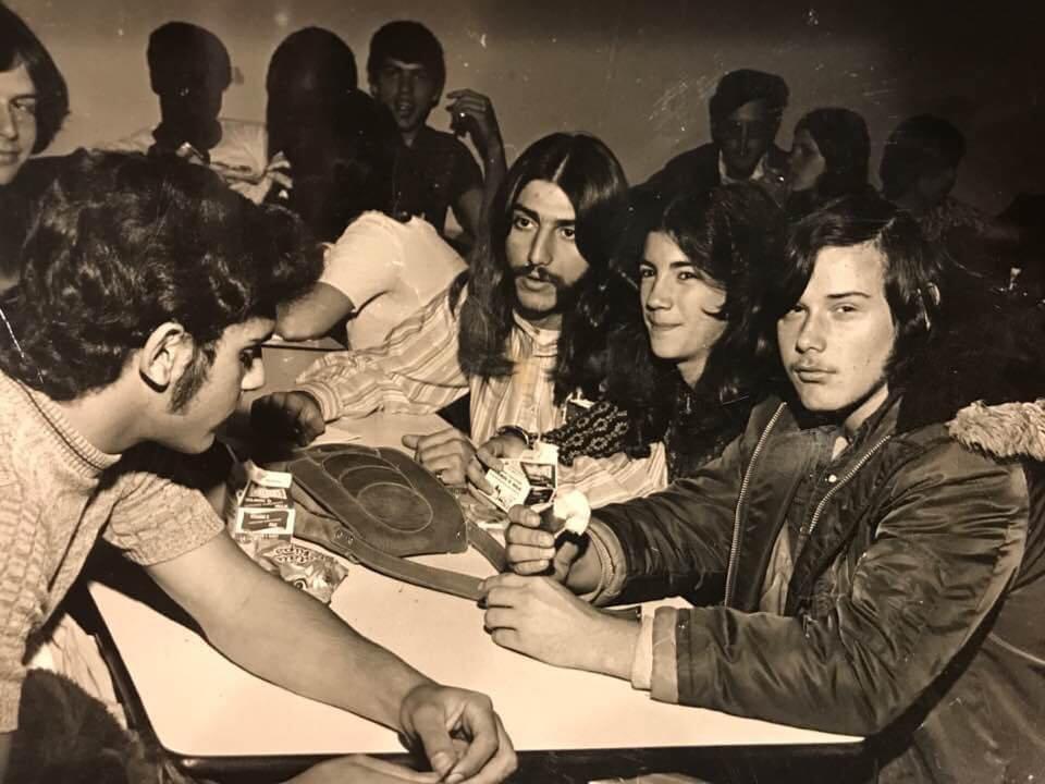 A black-and-white photo of a group of young men sitting around a table playing cards. Some are looking at the camera, while others are focused on the game. The scene suggests a casual, social gathering.