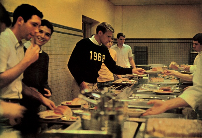 A group of young men stand in line at a cafeteria, receiving food from servers behind a counter. One man in front wears a dark sweater labeled "1968." The setting appears to be a school or college dining hall.