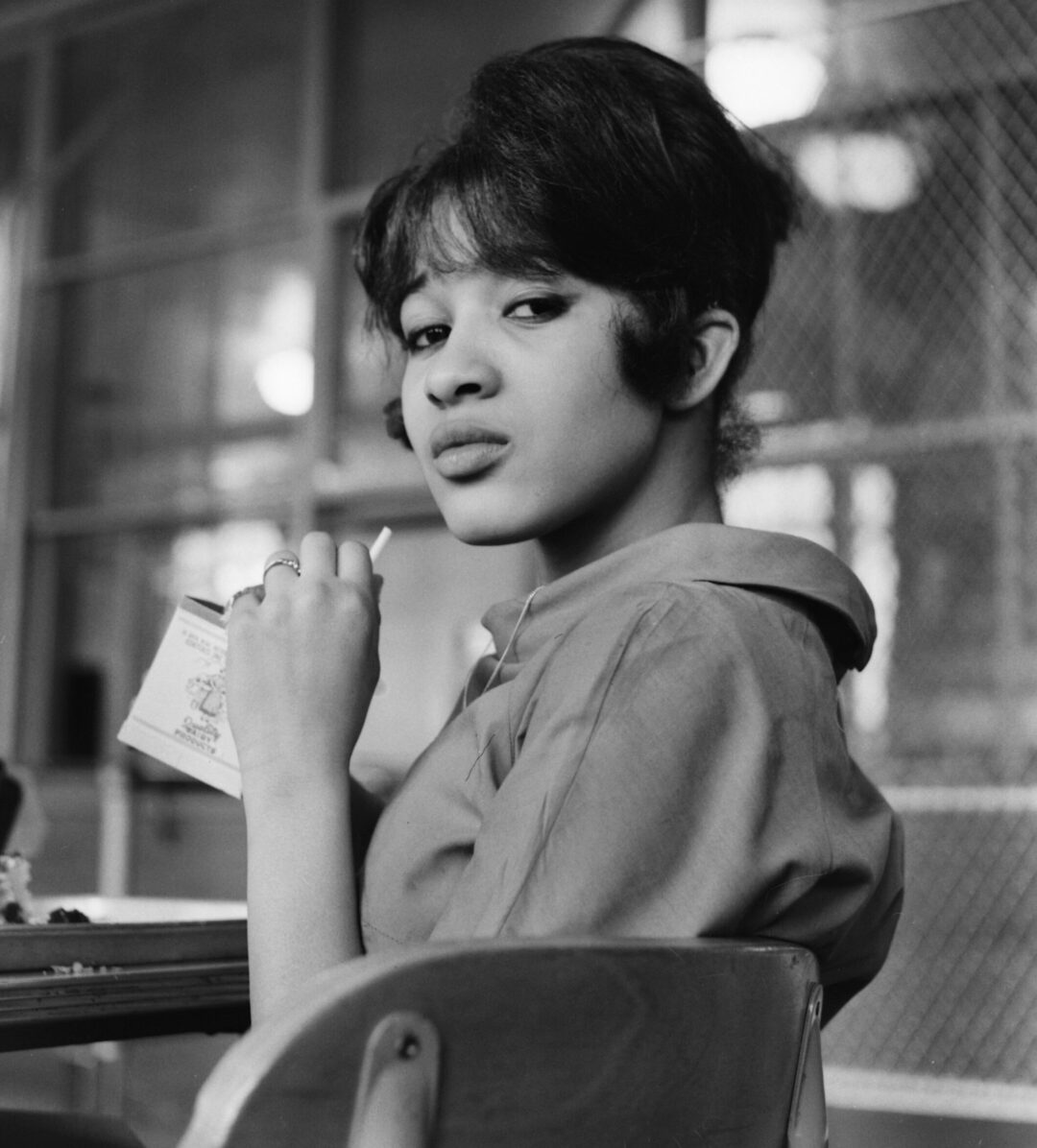 A young woman sits at a desk, holding a small milk carton and a straw. She has styled hair, wears a collared shirt, and looks slightly over her shoulder with a thoughtful expression in a classroom setting.