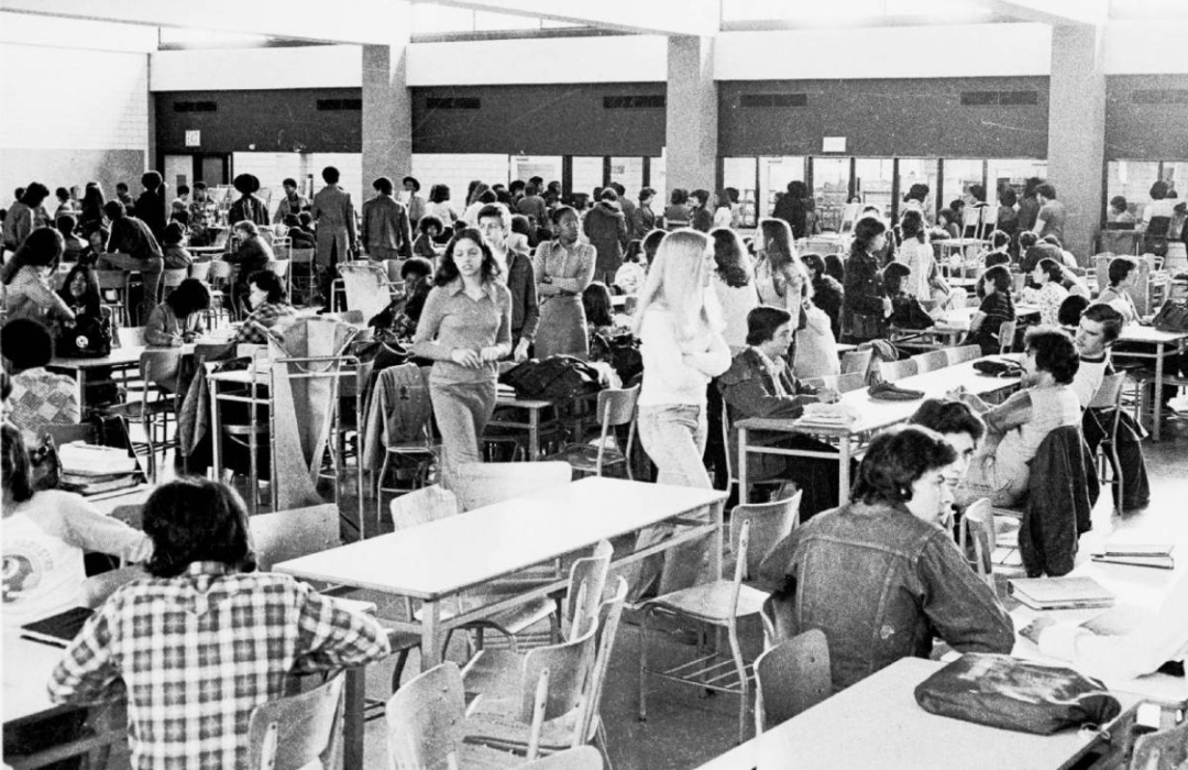Black-and-white photo of a busy school cafeteria filled with students sitting at tables, talking, eating, and walking around. The environment appears lively and social, with natural light coming through large windows.