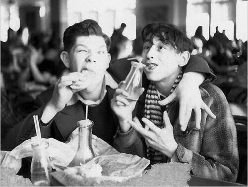 Two young men sit close together in a busy cafeteria, smiling and making playful faces as they eat sandwiches and drink from glass bottles with straws. One has his arm affectionately around the other.