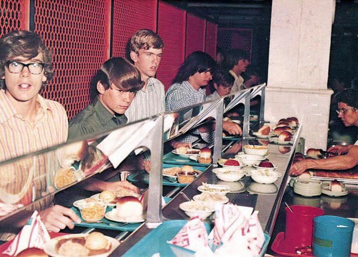 Teenagers stand in line at a cafeteria counter, holding trays as a staff member serves food. The counter is filled with plates of burgers, bowls, and wrapped items. The scene appears to be from the 1970s.