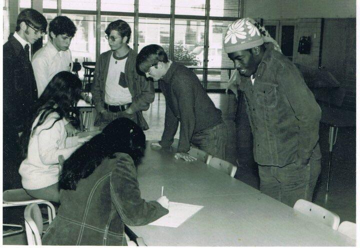 A black and white photo of four young men standing at a table while two women seated on the other side write on papers, with large windows in the background.