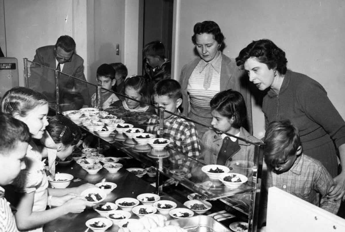 Black and white photo of children in a cafeteria line receiving food from behind a glass partition, with two women supervising and a man in the background. The children appear to be in elementary school.