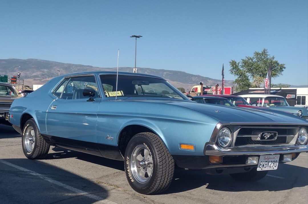 A light blue classic Ford Mustang car is parked at an outdoor car show on a sunny day, with mountains and a clear blue sky in the background. Other cars and people are visible in the distance.