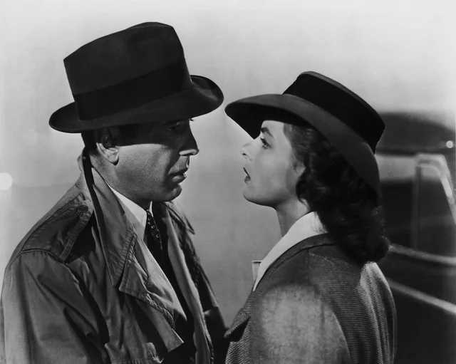 A man and a woman in vintage clothing and hats stand close together, facing each other with serious expressions, in a dramatic black-and-white scene suggesting classic film noir style.