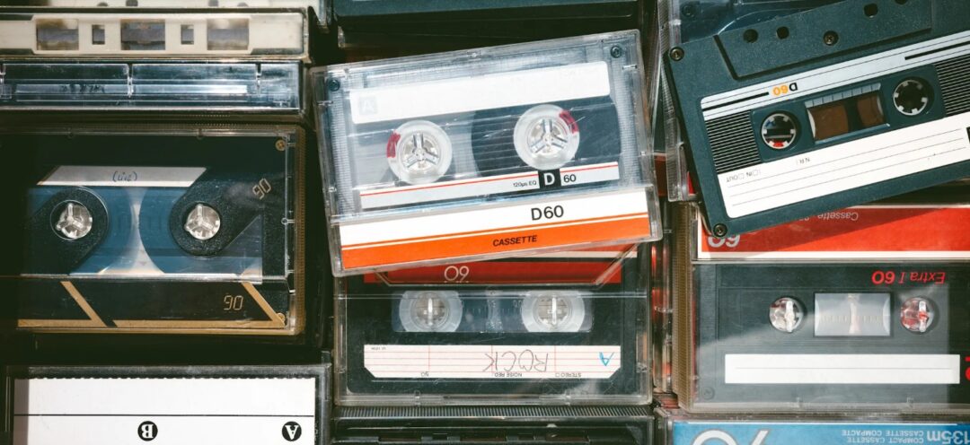 A close-up view of several vintage audio cassette tapes, some in cases, stacked and arranged in various orientations. The tapes have labels with different numbers and colors, showing signs of age and use.