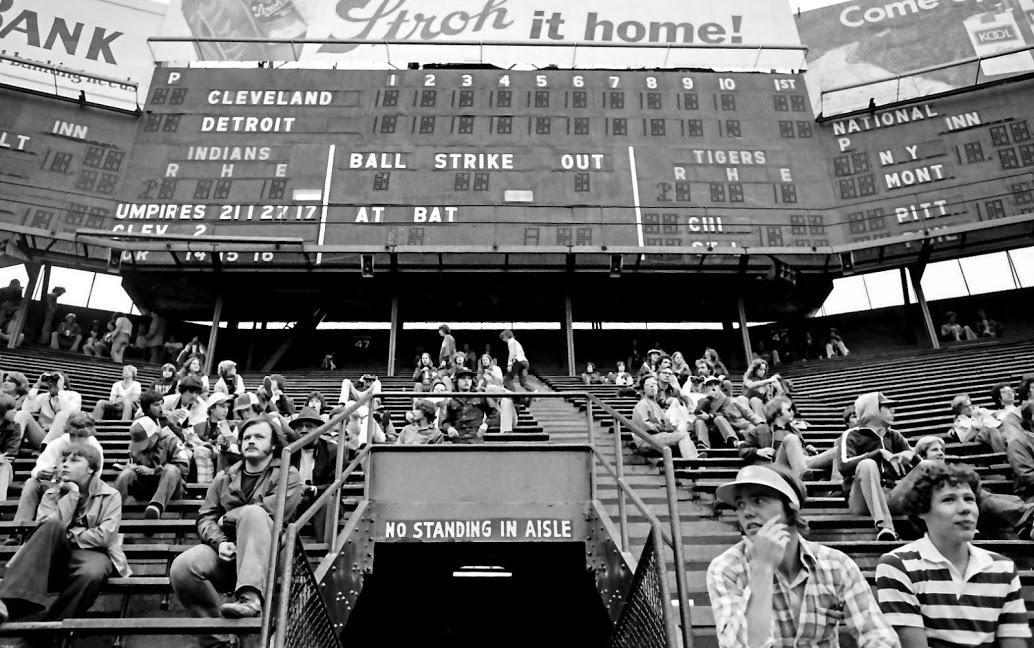 Black and white photo of a baseball stadium with scattered spectators sitting in bleachers beneath a large scoreboard showing the game between Cleveland Indians and Detroit Tigers. A sign reads "No Standing In Aisle.
