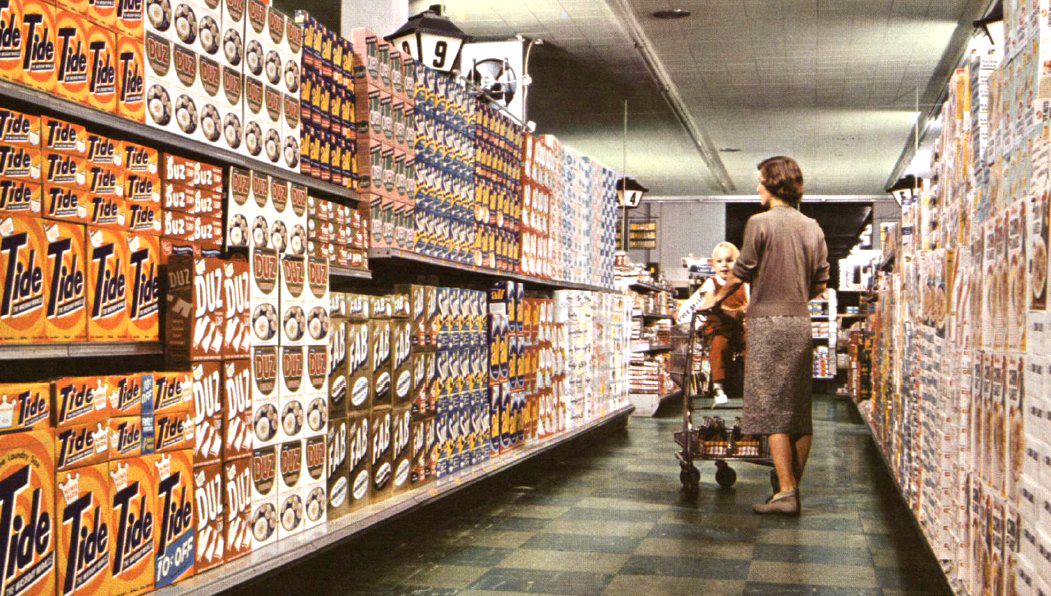 A woman pushes a shopping cart with a child in a grocery store aisle lined with shelves full of laundry detergent boxes, including Tide, under fluorescent lights.