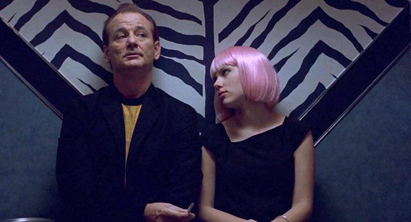 A man in a dark jacket sits next to a young woman with a short pink wig. They are in front of a zebra-striped wall, and the woman looks at the man while he looks away, both appearing thoughtful.