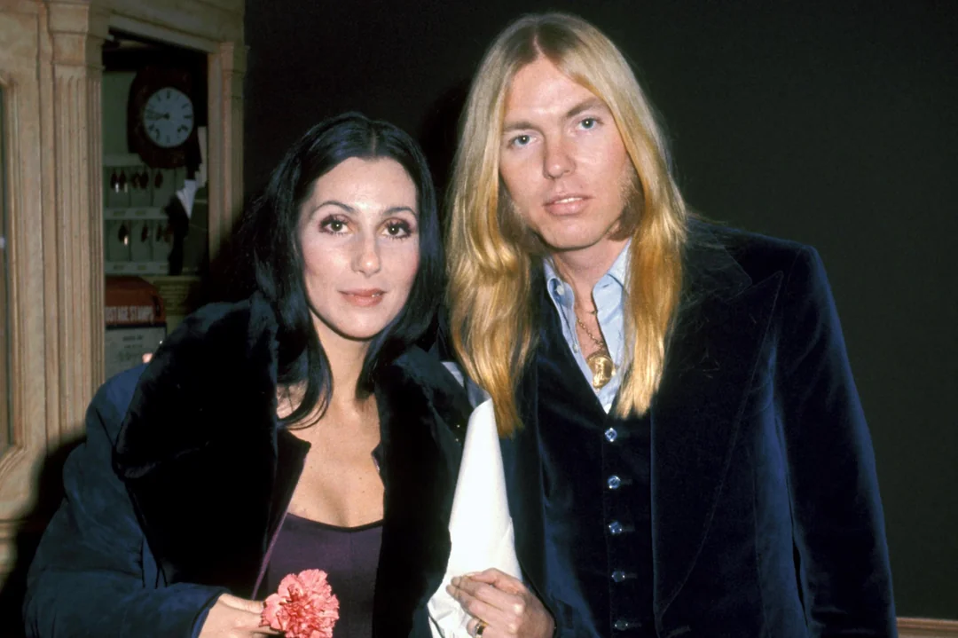 Two people stand close together, looking at the camera. The woman has long dark hair and wears a dark jacket, holding a pink flower. The man has long blond hair and wears a dark suit jacket over a light shirt.
