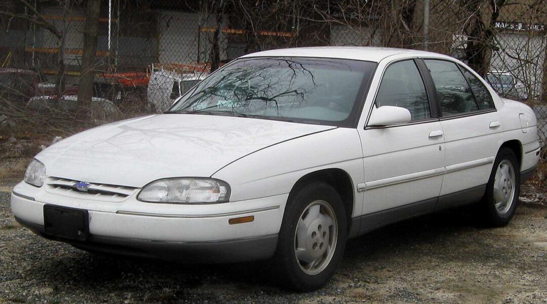 A white, four-door sedan car parked on a gravel surface with trees and a chain-link fence in the background. The car appears to be from the 1990s.