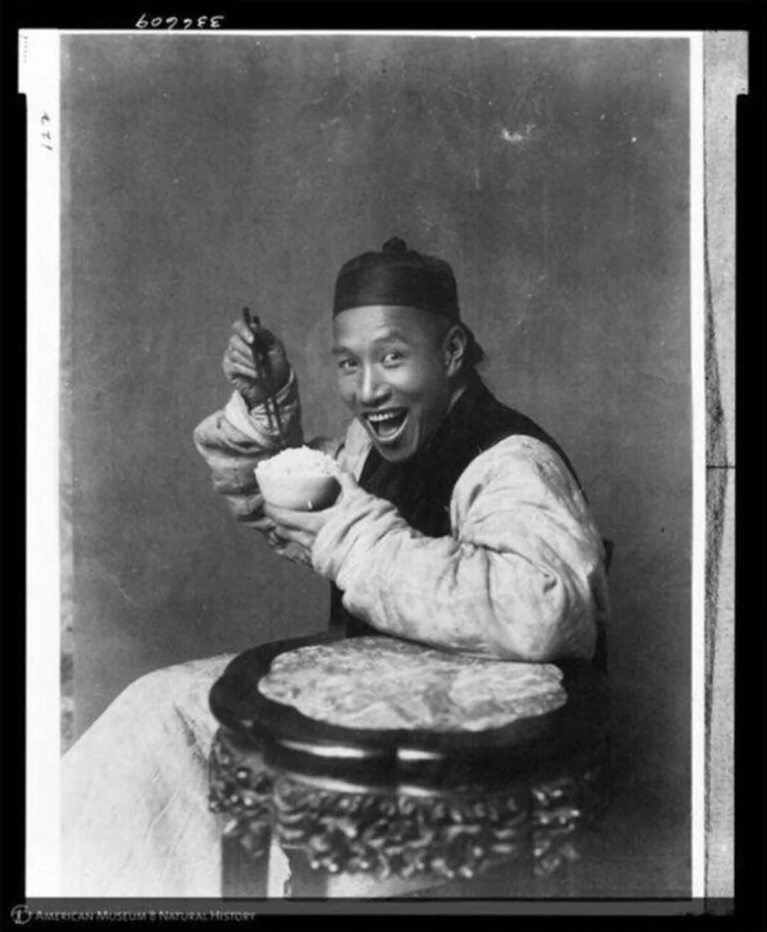 A man wearing traditional clothing and a cap sits on a carved stool, smiling broadly while holding chopsticks and a bowl, as if mid-bite. The background is plain and the photo is in black and white.