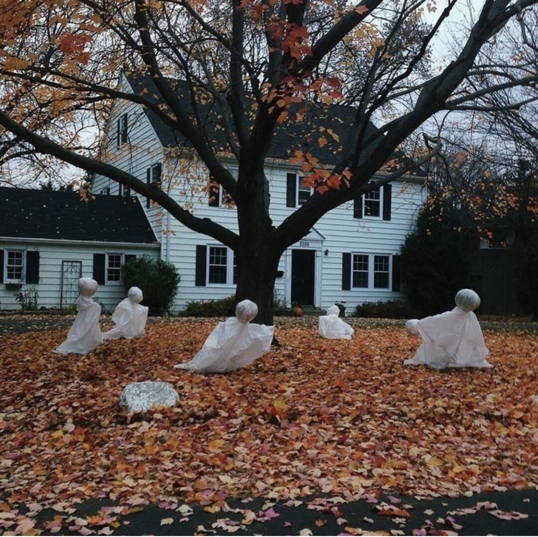 A yard covered in fallen autumn leaves features ghost decorations made from white sheets around a large tree, with a white house in the background.