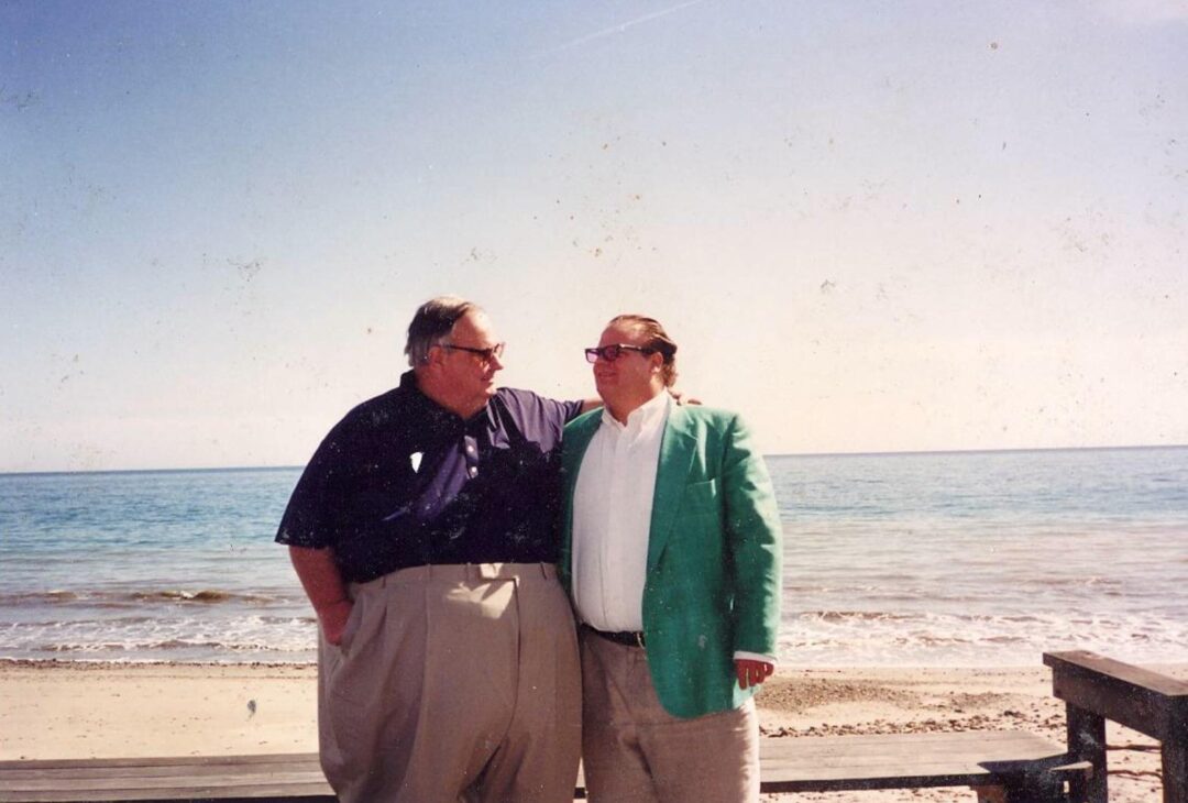 Two men stand close together on a beach, looking at each other and smiling. One wears a dark shirt and beige pants, the other wears a green blazer and white shirt. The ocean and clear sky are in the background.