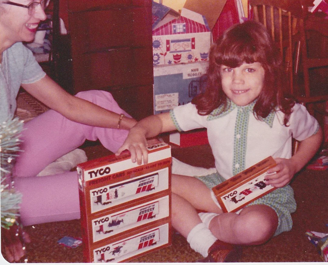 A young girl in a green-trimmed dress smiles while holding TYCO toy train boxes, sitting on the floor next to a woman. Wrapped gifts and a Christmas tree are visible in the background.