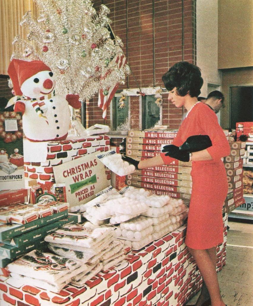 A woman in a red dress shops for Christmas decorations at a store display featuring a snowman, an artificial silver Christmas tree, boxes labeled "A BIG SELECTION," and bags of artificial snow.