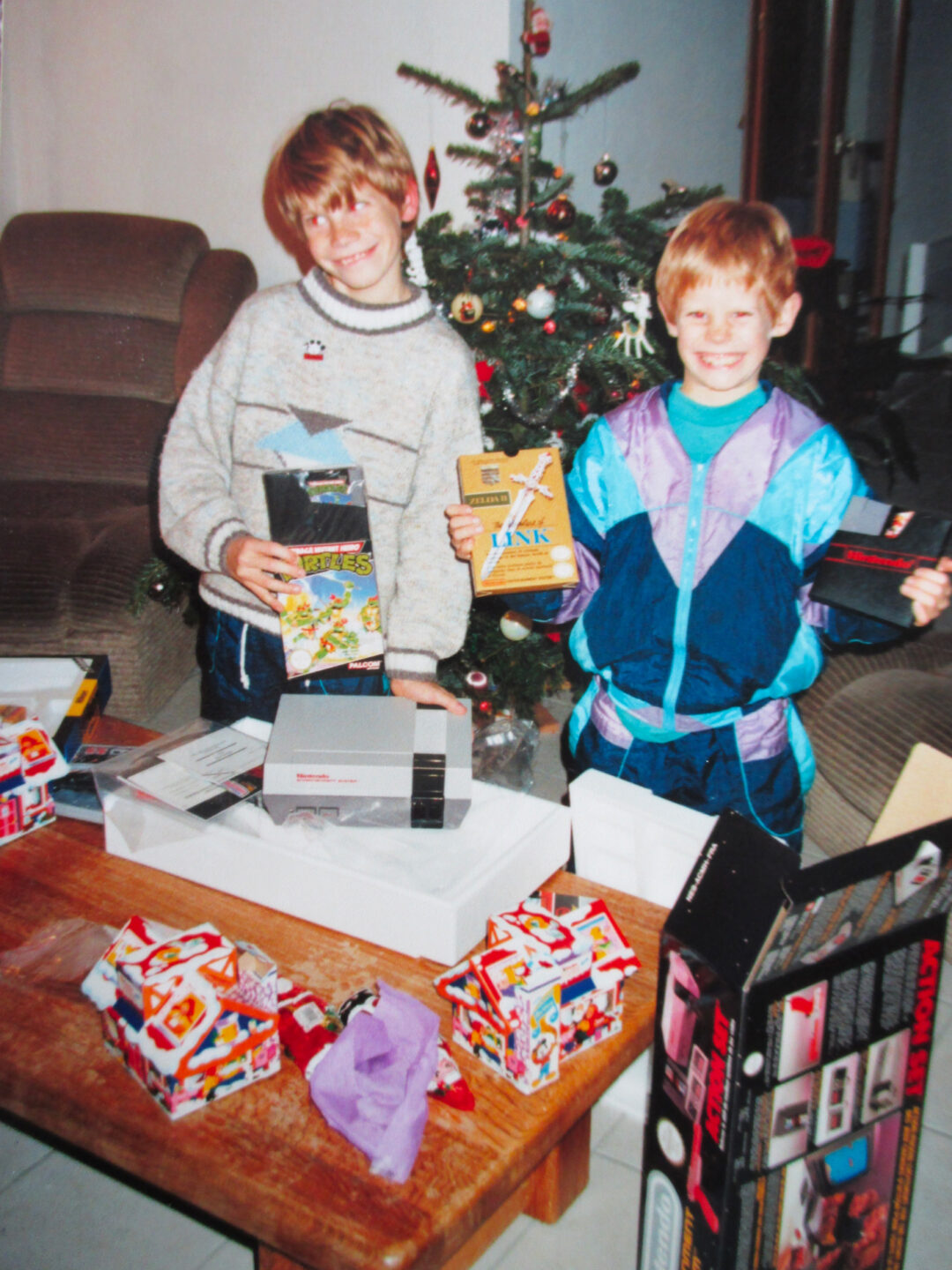 Two smiling children stand near a Christmas tree, holding video game cartridges and boxes. A Nintendo Entertainment System and wrapped gifts are on the table in front of them. The room is cozy with a brown couch in the background.