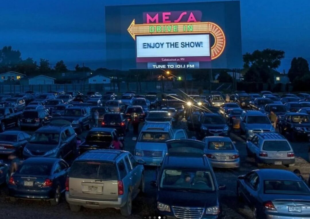 Cars are parked facing a large outdoor movie screen at dusk, displaying "MESA DRIVE IN" and "ENJOY THE SHOW!" People sit in their vehicles at the drive-in theater, with houses and trees in the background.