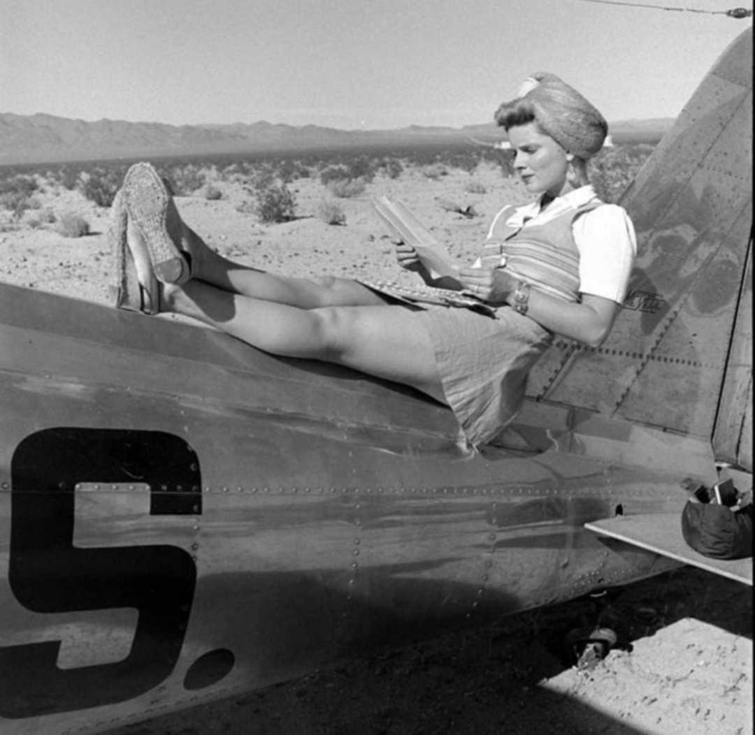 A woman in vintage clothing relaxes on the tail of an airplane, reading a book with her legs stretched out. The scene is set in a desert landscape with mountains in the distance under a clear sky.