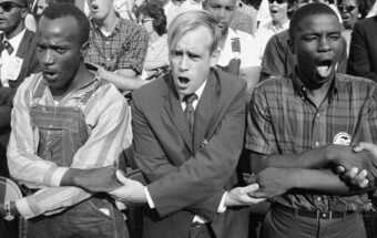 A diverse group of young men standing arm-in-arm, singing or chanting passionately during a civil rights demonstration, with people gathered closely behind them.