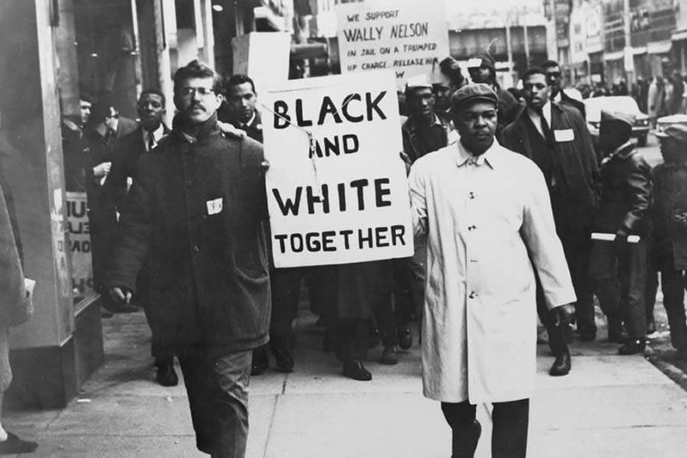 A group of civil rights protesters march down a city street. Two men at the front hold a sign that reads "BLACK AND WHITE TOGETHER." People march behind them, some carrying additional protest signs.