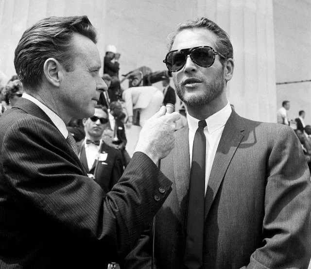 A man in a suit and sunglasses is being interviewed by another man holding a microphone. They stand outdoors with a crowd and large stone columns in the background. The scene appears to be from the mid-20th century.