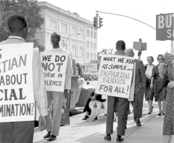 Black men participate in a peaceful protest on a city sidewalk, wearing signs demanding impartial service and an end to racial discrimination, while pedestrians walk past and observe.