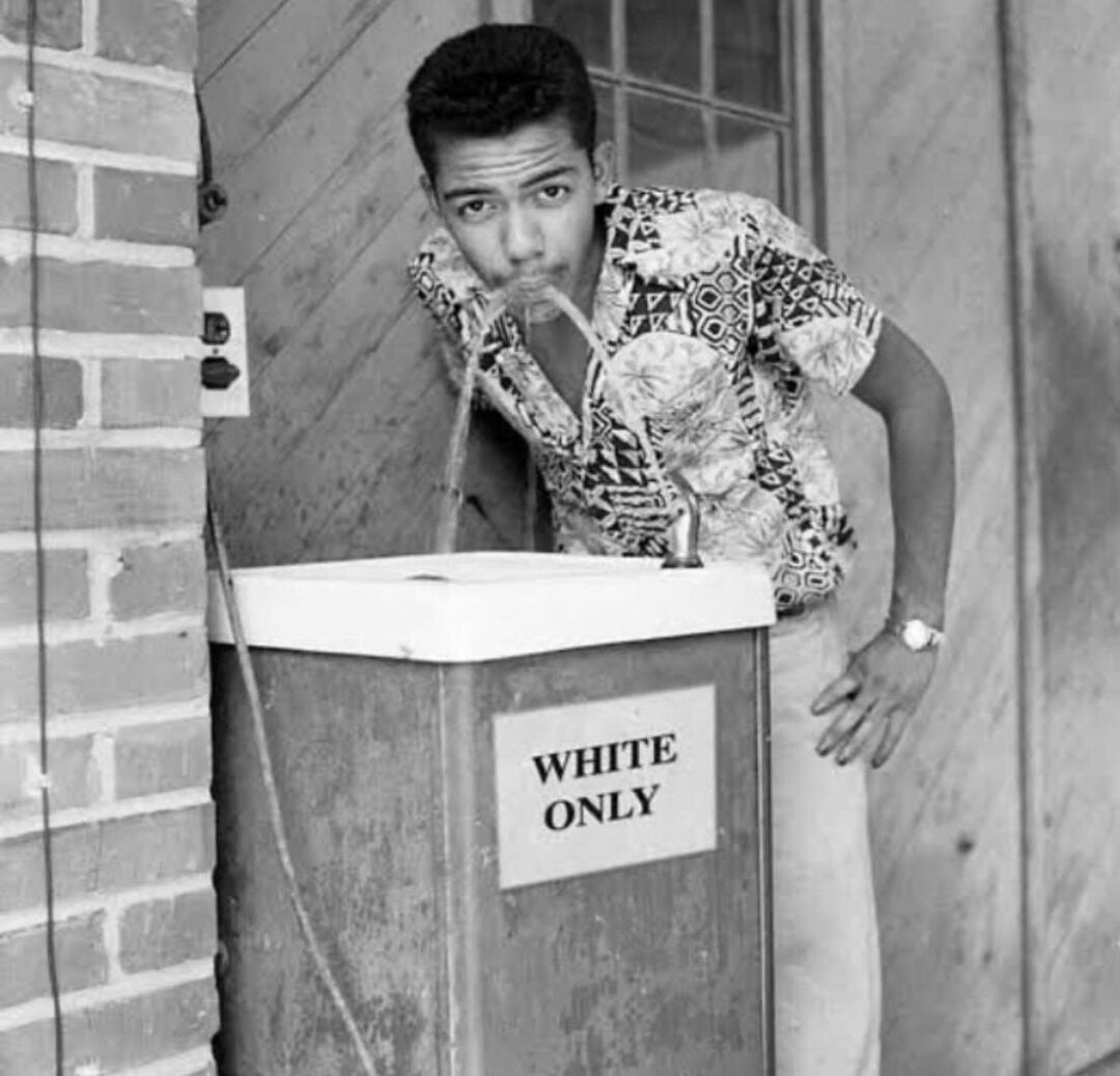 A young Black man drinks from a water fountain labeled "WHITE ONLY" in bold letters, highlighting segregation during the Jim Crow era. The scene is in black and white, set against a brick and wood wall.