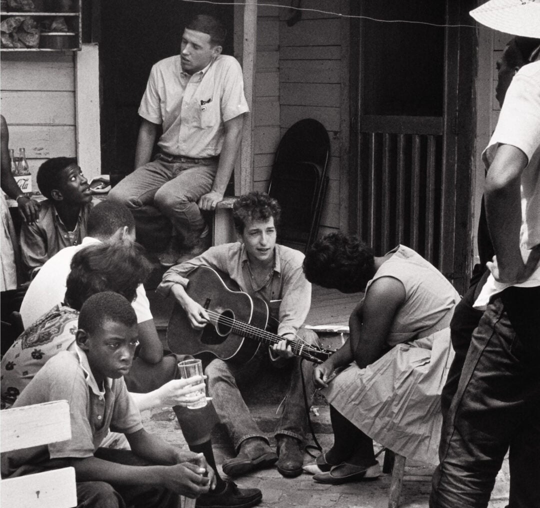 A black-and-white photo of a group of people gathered outside a house. One person sits on the ground playing guitar, others sit or stand around, listening and talking. The scene appears casual and relaxed.