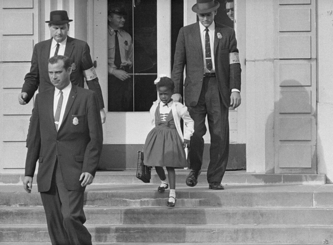 A young Black girl in a dress carries a school bag while being escorted down school steps by four suited, white men with badges and armbands, likely federal agents, in a historic, black-and-white photograph.