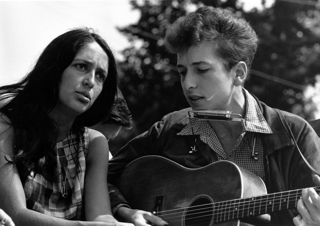 A woman and a man sit closely together outdoors. The man strums a guitar and wears a harmonica holder, while the woman looks ahead and appears to be singing or speaking. Trees and blurred figures are in the background.