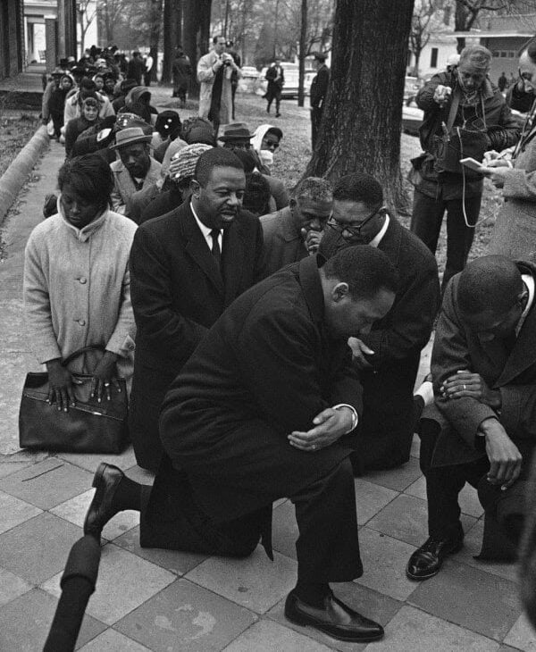 A group of civil rights activists, including men and women, kneel in prayer or protest on a sidewalk, some with bowed heads, while others stand and observe in the background. Trees and houses line the street.