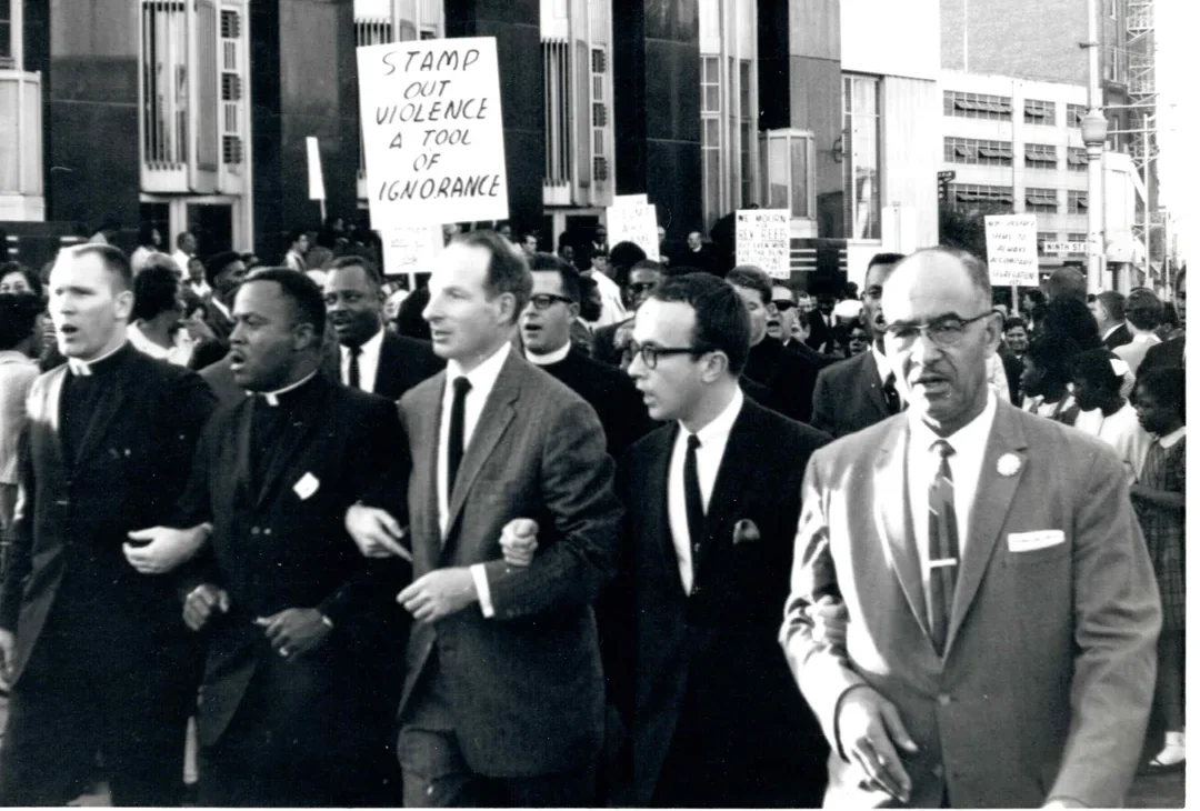 A group of civil rights activists link arms at a protest march in front of a building. One holds a sign reading “Stamp out violence, a tool of ignorance.” The photo is dated June 1965.