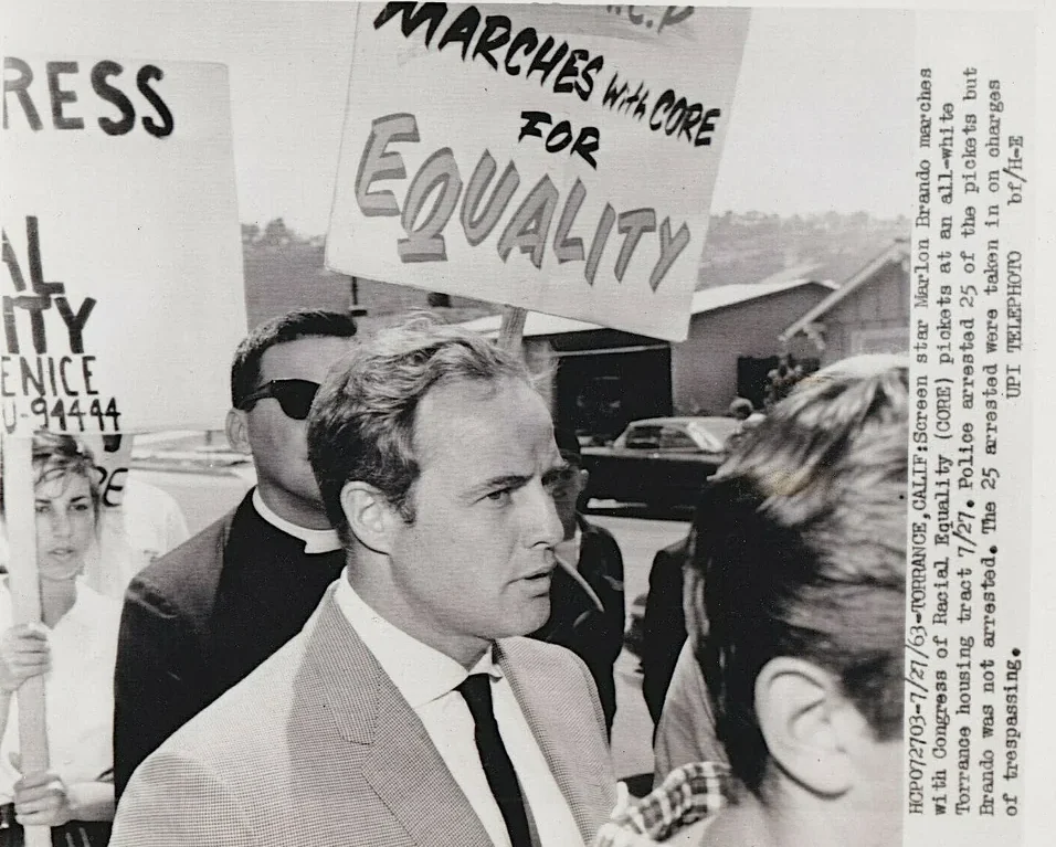 A man in a suit marches in a crowd holding signs that read "Marches with CORE for equality" and partial text "EQUALITY." People around him also carry signs during a civil rights protest.