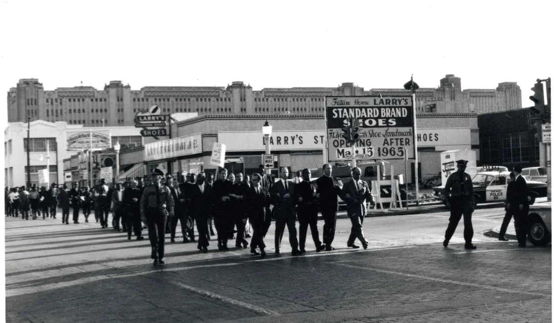 A large group of people, mostly men in suits, march together down a city street in front of storefronts, including Larry’s Shoes. The photo is black and white and dated June 1965.