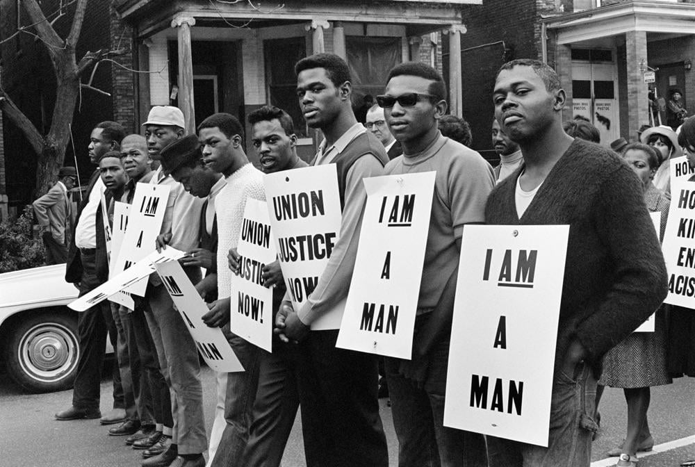 A group of Black men stand in a line on a city street holding protest signs that read “I AM A MAN” and “UNION JUSTICE NOW” during a civil rights demonstration in front of brick houses.