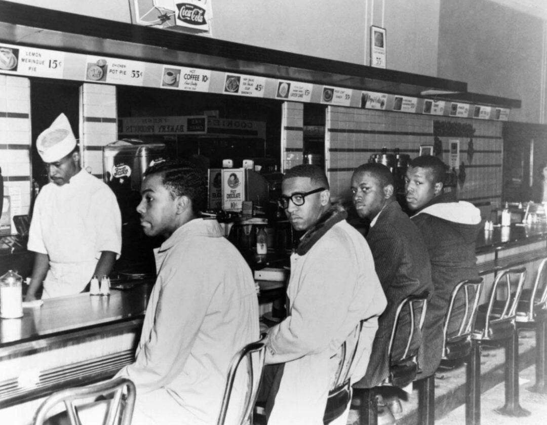 Four young Black men sit at a lunch counter, looking forward, while a worker behind the counter stands nearby. The setting appears to be a mid-20th-century diner, with menu boards visible above.